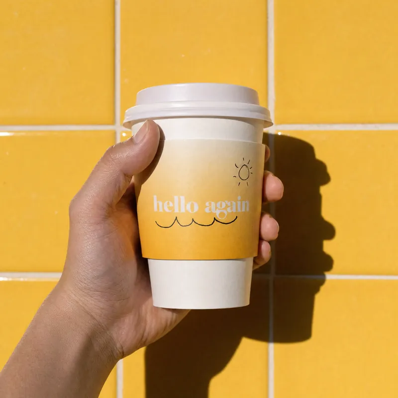 A hand holding a Hello Again cup against yellow tiles in hard sun