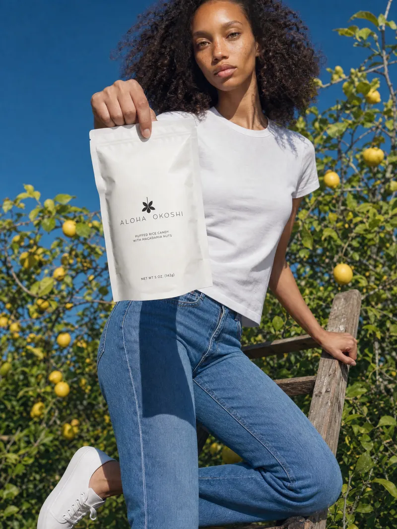 Noa on an orchard ladder holding an Aloha Okoshi bag toward the camera by its top edge
