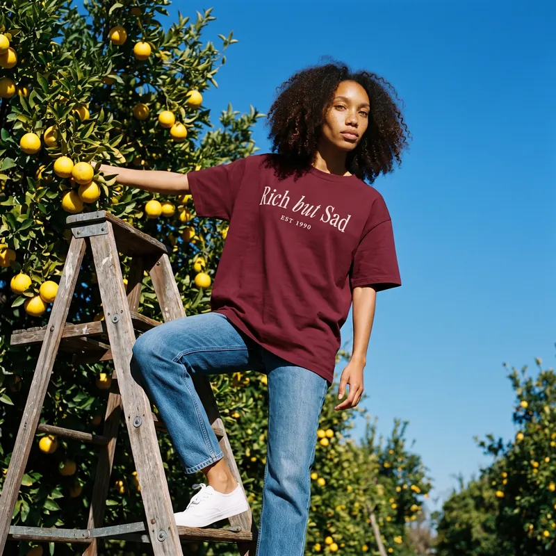 Noa on an orchard ladder in the burgundy Rich But Sad Monaco tee