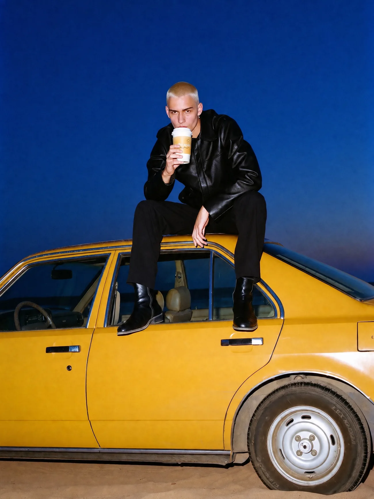 A man in a leather jacket sits on a yellow car at dusk holding a Hello Again cup