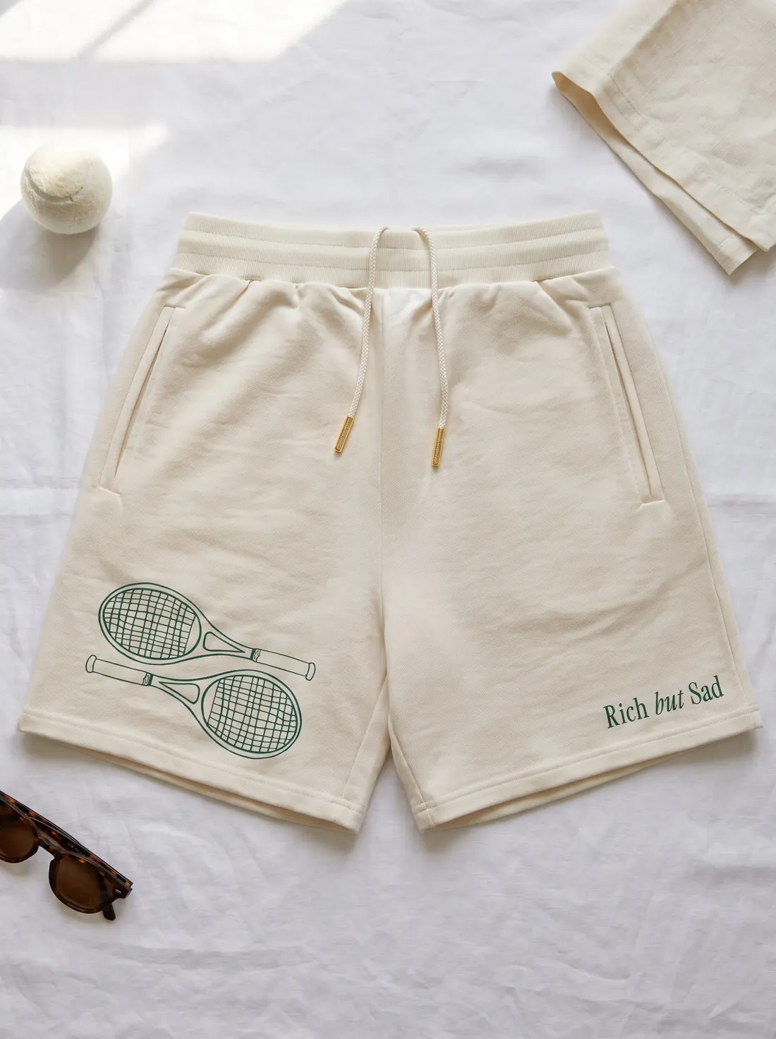 Cream Rich But Sad shorts with a racket print laid flat on white linen